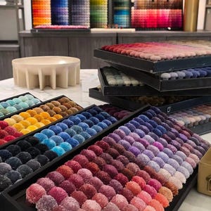 Pu&ograve; includere: Diversi vassoi rettangolari riempiti con file di piccoli pompon in tessuto rotondi e testurizzati in un ampio spettro di colori, tra cui sfumature di rosso, rosa, viola, blu, giallo e nero, disposti in gradienti per la selezione.