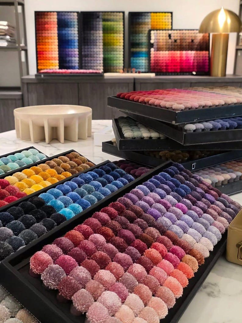 Pu&ograve; includere: Diversi vassoi rettangolari riempiti con file di piccoli pompon in tessuto rotondi e testurizzati in un ampio spettro di colori, tra cui sfumature di rosso, rosa, viola, blu, giallo e nero, disposti in gradienti per la selezione.