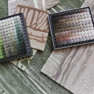Pu&ograve; includere: Due cartelle colori rettangolari con una griglia di piccoli campioni di tessuto trapuntato circolare in varie tonalit&agrave; di verde e marrone, disposte sopra diversi campioni di tessuto testurizzato con motivi astratti in beige, grigio e verde.