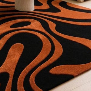 Pu&ograve; includere: Un tappeto rettangolare caratterizzato da un motivo astratto ad alto contrasto con forme organiche fluide in nero profondo e arancione bruciato. Il design &egrave; composto da spesse linee ondulate che creano un senso di movimento sulla superficie.