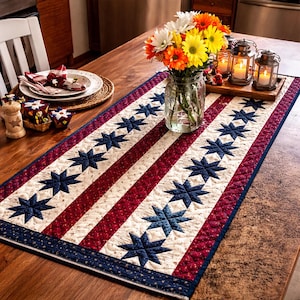 May include: A quilted table runner with a patriotic design. It features alternating red and white stripes with blue star accents. The runner is on a wooden table, with a vase of flowers and candles.