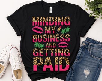 Può includere: T-shirt nero con la scritta "MINDING MY BUSINESS AND GETTING PAID" in rosa e stampa leopardata. Il design include rossetto, banconote e labbra rosa. Sono visibili scarpe da ginnastica bianche e jeans blu.