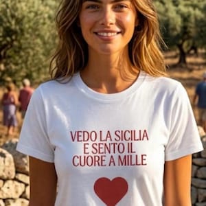 Può includere: T-shirt bianco con la scritta italiana "VEDO LA SICILIA E SENTO IL CUORE A MILLE" in rosso e un cuore rosso. La persona indossa la maglietta all'aperto.