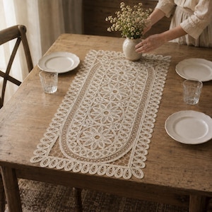May include: A rectangular beige lace table runner featuring a repeating floral pattern with circular motifs and a scalloped edge. The runner is centered on a wooden table with white ceramic plates and clear glass tumblers placed around it.