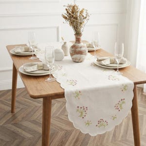 Puede incluir: Una mesa de comedor de madera preparada para una comida. Un camino de mesa blanco con diseños florales bordados está centrado en la mesa. Platos, servilletas y copas de champán están dispuestos para cada cubierto. Un jarrón con flores secas está en el centro.