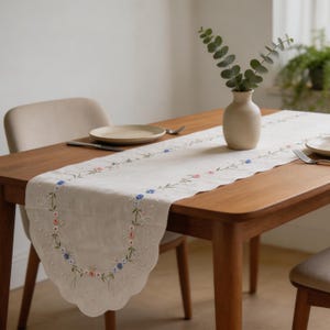 May include: A cream-coloured embroidered table runner with a scalloped edge. The runner features a floral design in pink and blue, with green stems and leaves. The runner is placed on a wooden table, with a vase of greenery and place settings visible.