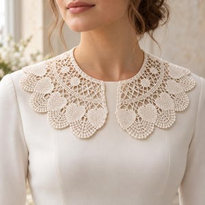 May include: A detachable off-white crochet collar featuring an intricate openwork pattern with repeating heart-shaped motifs along the scalloped edge. The collar is worn over a plain, long-sleeved, off-white garment with a round neckline.