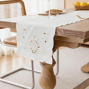 Può includere: Runner da tavolo bianco ricamato con bordo smerlato, con un motivo floreale centrale e dettagli traforati. Il runner è posto su un tavolo di legno, creando un'elegante ambientazione.