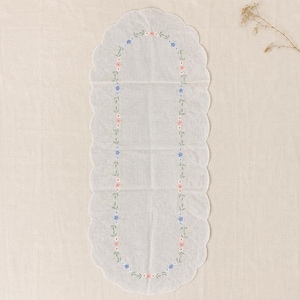 May include: An oval-shaped white table runner featuring a scalloped edge and a delicate embroidered border of small pink and blue flowers with green stems arranged in a continuous loop pattern across the center of the sheer fabric surface.