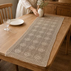 May include: A rectangular beige crochet table runner featuring a repeating floral motif arranged in a diamond grid pattern, finished with a delicate scalloped edge border. The intricate needlework displays a consistent openwork design.