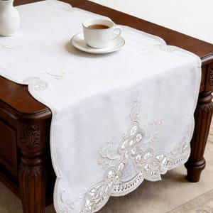 May include: White embroidered table runner with a scalloped edge, featuring delicate floral and geometric patterns. A white teacup and saucer with tea sit on the runner, placed on a dark wooden table. A white vase is also visible.