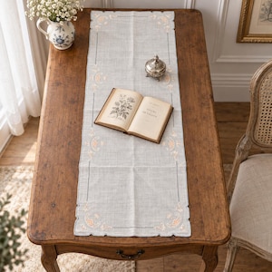 May include: A rectangular light gray table runner featuring delicate peach and white floral embroidery at the corners and edges. The runner is centered on a wooden table, with an open book titled Fleurs de France and a small metal trinket box.