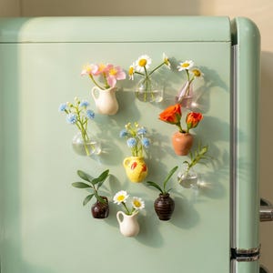 Könnte beinhalten: Eine Ansammlung kleiner, farbenfroher Keramikvasen und Glasbehälter, die jeweils eine andere Art von Blume enthalten, angeordnet auf einer hellgrünen Kühlschranktür. Die Vasen haben verschiedene Formen und Farben, darunter Weiß, Gelb und Braun.