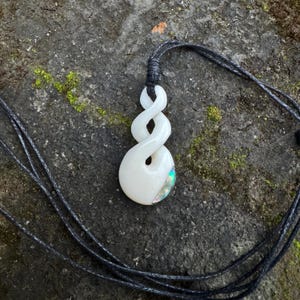 Puede incluir: Un colgante blanco con un diseño de doble bucle retorcido suspendido de un cordón gris oscuro. El colgante presenta una superficie lisa y pulida con una pequeña incrustación iridiscente en la parte inferior derecha que refleja tonos de luz verdes y azules.