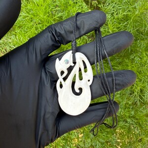 May include: A white, carved bone pendant featuring an intricate, swirling openwork design with a small, iridescent abalone shell inlay. The pendant is suspended from a thin, black braided cord, creating a contrast against the smooth, light material.