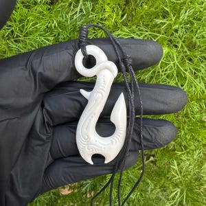 May include: A white, carved fish hook pendant featuring intricate, swirling patterns on its surface. The pendant is suspended from a thin, black cord with adjustable sliding knots, creating a functional and detailed piece of jewelry.