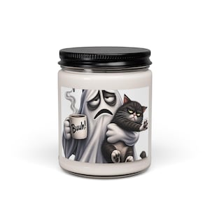 Könnte beinhalten: Eine weiße Kerze in einem Glas mit schwarzem Deckel, versehen mit einem Etikett, das einen Cartoon-Geist zeigt, der eine dampfende Tasse mit der Aufschrift Buuh! hält und eine mürrische schwarze Katze trägt. Der Geist wirkt müde, die Katze streng.