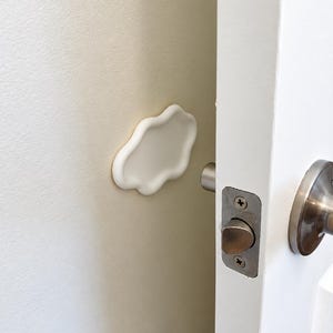 May include: A white cloud-shaped wall protector mounted on a beige wall, positioned to cushion the impact of a metal door handle. The door is white with a silver-toned latch mechanism and a circular handle plate visible on the right side.