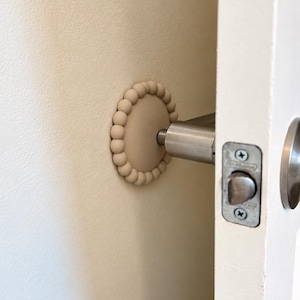 May include: A beige circular wall protector featuring a decorative beaded border. The protector is mounted on a wall behind a silver metal door handle, serving as a buffer to prevent the handle from making direct contact with the wall surface.