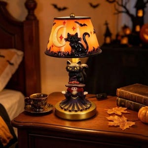 May include: A table lamp featuring an orange shade decorated with a black cat, crescent moon, and bats. The base is a sculpted figure of a witch wearing a purple dress with a black cat perched on her hat, all set on a circular gold-toned stand.