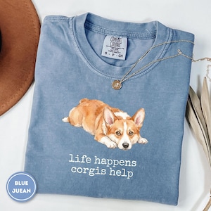 May include: A blue short-sleeved t-shirt featuring a watercolor-style illustration of a brown and white Corgi dog lying down. Below the dog, the text reads "life happens corgis help" in white letters. The shirt has a Comfort Colors brand label.
