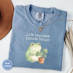 May include: A blue short-sleeve t-shirt featuring a graphic of a light green frog holding a small potted plant. The text "life happens plants helps" is printed in white above the frog. The shirt has a crew neckline and a Comfort Colors label.