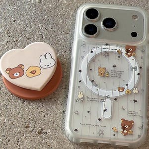 Op de afbeelding: Transparante telefoonhoes met een cartoon beer- en konijnontwerp, plus een bijpassende hartvormige telefoonhouder. De hoes heeft een magnetische ring en een sterrenpatroon. De telefoonhouder is lichtbruin en perzik.