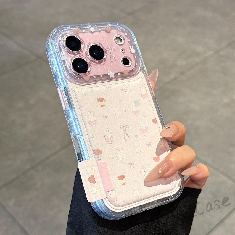 May include: Clear phone case with a pink interior featuring a pattern of cartoon bunnies, bows, and stars. The case has a clear, protective bumper and a pink accent at the bottom. The phone case is held by a person.