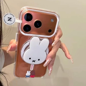K&ouml;nnte beinhalten: Transparente Handyh&uuml;lle mit einem wei&szlig;en Miffy-Hasen-Design. Die H&uuml;lle hat eine ros&eacute;goldene R&uuml;ckseite und einen Pop-up-Griff. Der Text "Miffy x Case" ist ebenfalls vorhanden.