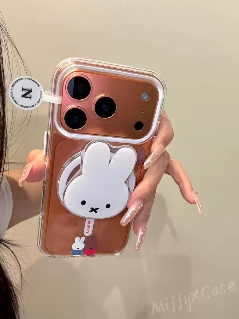 K&ouml;nnte beinhalten: Transparente Handyh&uuml;lle mit einem wei&szlig;en Miffy-Hasen-Design. Die H&uuml;lle hat eine ros&eacute;goldene R&uuml;ckseite und einen Pop-up-Griff. Der Text "Miffy x Case" ist ebenfalls vorhanden.