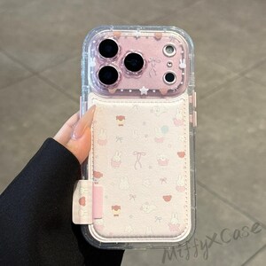 May include: Clear phone case with a pink interior and a white card holder. The card holder features a repeating pattern of cartoon bunnies, bows, and hearts. The case has a clear border and a pink camera lens protector.