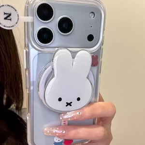 K&ouml;nnte beinhalten: Transparente Handyh&uuml;lle mit einem wei&szlig;en Miffy-Hasen-Design und einem passenden Pop-up-Griff. Die H&uuml;lle hat eine transparente R&uuml;ckseite und einen wei&szlig;en hasenf&ouml;rmigen Griff. Auf der Seite ist auch ein kleiner rosa Hase zu sehen.