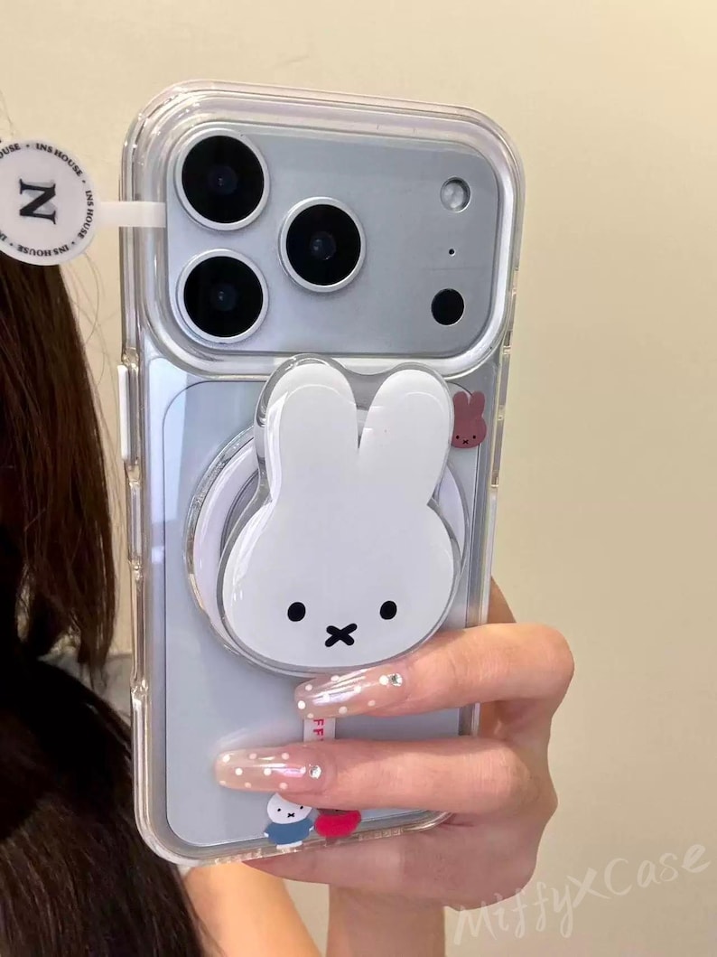 K&ouml;nnte beinhalten: Transparente Handyh&uuml;lle mit einem wei&szlig;en Miffy-Hasen-Design und einem passenden Pop-up-Griff. Die H&uuml;lle hat eine transparente R&uuml;ckseite und einen wei&szlig;en hasenf&ouml;rmigen Griff. Auf der Seite ist auch ein kleiner rosa Hase zu sehen.