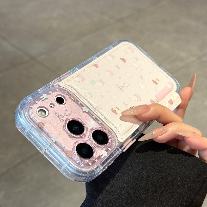 May include: A clear phone case with a pink interior and a white cardholder. The cardholder features a repeating pattern of cartoon characters in pink. The case has a clear, glittery border.