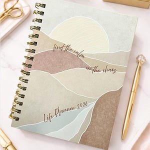 May include: A spiral-bound Life Planner 2026 with a serene landscape design in muted tones of beige, brown, and blue. The cover features the text "find the calm in the chaos." A gold pen rests beside the planner.
