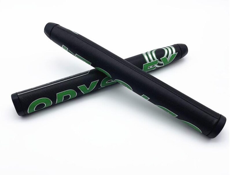 May include: Two black golf putter grips with green lettering and accents are displayed against a white background. The grips are cylindrical and textured, with the word "ODYSSEY" visible. One grip is positioned over the other.