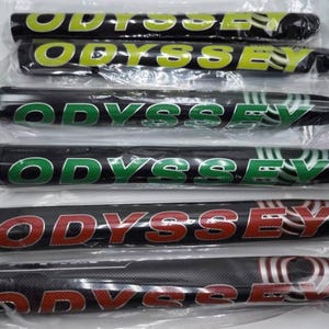 K&ouml;nnte beinhalten: Mehrere schwarze Golfschl&auml;gergriffe mit dem Wort "ODYSSEY" in verschiedenen Farben, darunter Gr&uuml;n, Gelb und Rot. Die Griffe sind in durchsichtigem Kunststoff verpackt.