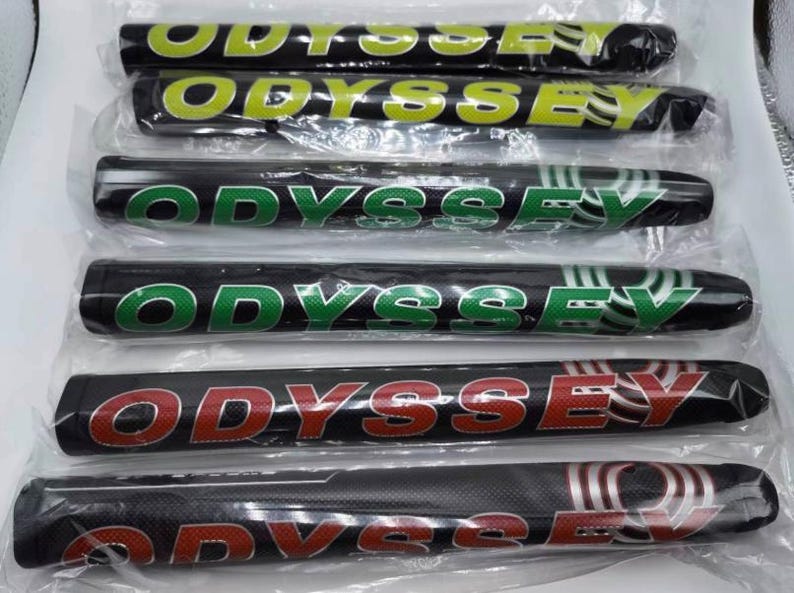 K&ouml;nnte beinhalten: Mehrere schwarze Golfschl&auml;gergriffe mit dem Wort "ODYSSEY" in verschiedenen Farben, darunter Gr&uuml;n, Gelb und Rot. Die Griffe sind in durchsichtigem Kunststoff verpackt.