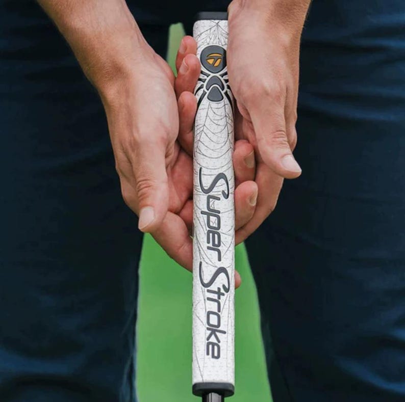 Pu&ograve; includere: Impugnatura per putter da golf bianca con la scritta "Super Stroke" in nero. L'impugnatura presenta un design a ragnatela e una grafica di ragno nera e grigia. L'impugnatura &egrave; tenuta da due mani.