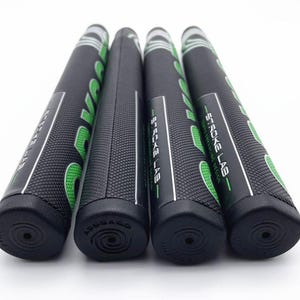 May include: Four black golf club grips with the text "STROKE LAB" in white and green. The grips are cylindrical with a textured surface and a black end cap. The grips are arranged in a row against a white background.