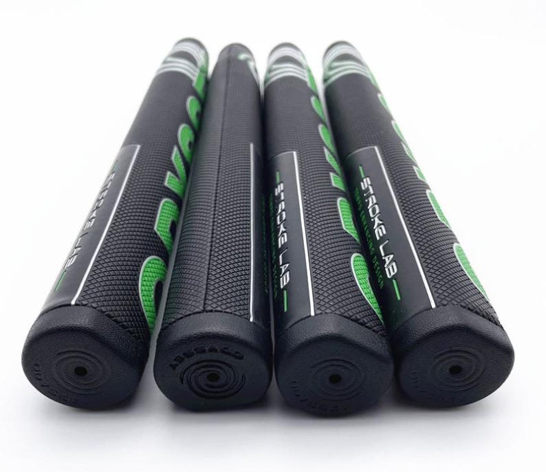 May include: Four black golf club grips with the text "STROKE LAB" in white and green. The grips are cylindrical with a textured surface and a black end cap. The grips are arranged in a row against a white background.
