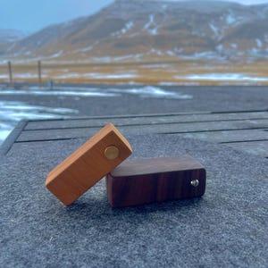 Peut inclure: Deux boîtes rectangulaires en bois, une marron clair et une marron foncé, avec des fermetures circulaires en laiton ou argent. Les boîtes reposent sur une surface grise, avec un arrière-plan de montagnes et un ciel nuageux.