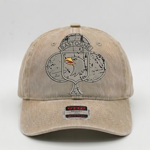 Puede incluir: Gorra de béisbol beige con un emblema desgastado que presenta un águila y la palabra "BASTOGNE". La gorra tiene una visera curva y una correa ajustable. La etiqueta de la marca "OTTO" es visible.
