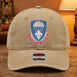 Puede incluir: Gorra beige con un parche que presenta un paracaídas, una flor de lis y las palabras "Supporting Freedom". La gorra tiene una visera curva y un ajuste relajado. La etiqueta dice "Dad Hat" y "Maximum Comfort".