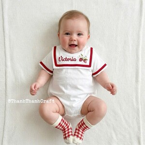 Op de afbeelding: Een baby in een witte romper met rode bies en de naam "Victoria" geborduurd op de kraag. De romper heeft een aardbeienontwerp en wordt gecombineerd met rood-witte geruite sokken. De baby ligt op een witte deken.