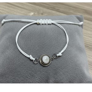 Peut inclure: Bracelet en macramé blanc avec un pendentif circulaire argenté. Le pendentif présente une pierre blanche et ronde en forme de coquillage. Le bracelet est présenté sur une surface grise.