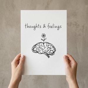 Puede incluir: Una impresión en papel blanco sostenida por manos, con las palabras "thoughts & feelings" en una fuente manuscrita. Debajo del texto hay un dibujo lineal negro de un cerebro con una flor que crece.