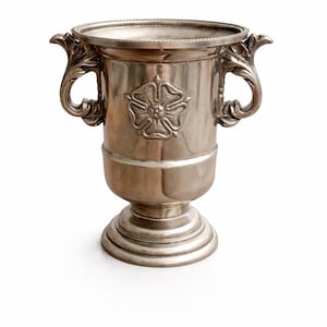 Puede incluir: Trofeo plateado con dos asas ornamentadas y un emblema floral. El trofeo tiene una parte superior ancha y abierta y una base de pedestal. El diseño es detallado y el metal tiene un acabado pulido. Probablemente se utiliza para premios o exhibiciones.