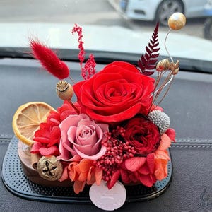 May include: A vibrant floral arrangement featuring a large red rose as the focal point, complemented by smaller pink and red blooms. Dried lemon slices and decorative elements add texture. The arrangement is set in a wooden base with a small, round, white label.