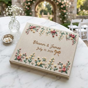 Può includere: Libro degli ospiti di nozze in lino beige con disegni floreali ricamati in rosa, giallo e verde. I nomi "Eleanor & James" e la data "10 luglio 2026" sono ricamati con filo dorato. È presente anche un piccolo fiocco ricamato.